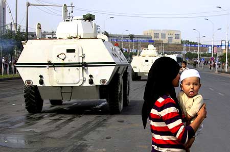 An ethnic Uighur woman carries her child across the road as Chinese armoured personnel carriers drive past