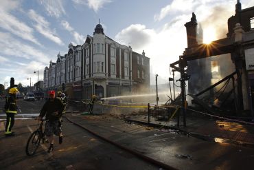 A cyclist passes firemen dowsing down buildings set alight during riots in Tottenham, north London.