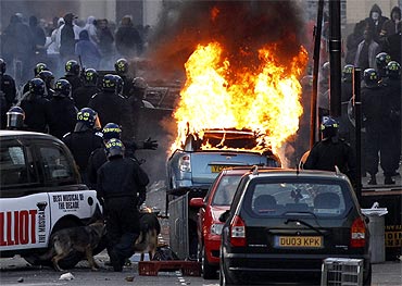 Police officers in riot gear block a road near a burning car on a street in Hackney, east London