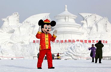 A man dressed as Mickey Mouse poses in front of a snow sculpture at a park