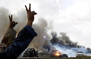 Rebel fighters gesture in front of burning vehicles belonging to Gaddafi's forces after an air strike by coalition forces along a road between Benghazi and Ajdabiyah in Libya