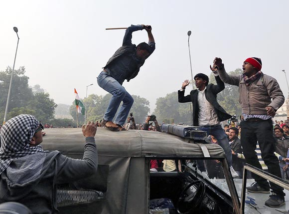 A demonstrator breaks the windshield of a police vehicle as others shout slogans in front of the India Gate