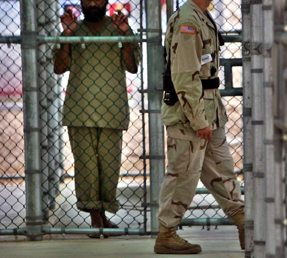 In this photo reviewed by US military officials, a detainee whose name, nationality, and facial identification are not permitted, holds onto a fence as a US military guard walks past the grounds of the maximum security prison at Camp 5 in the Guantanamo Bay