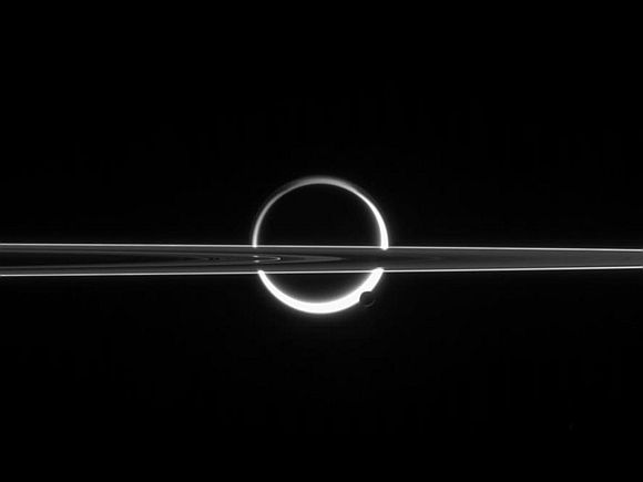 Saturn's rings cut across an eerie scene that is ruled by Titan's luminous crescent and globe-encircling haze, broken by the small moon Enceladus, whose icy jets are dimly visible at its south pole.