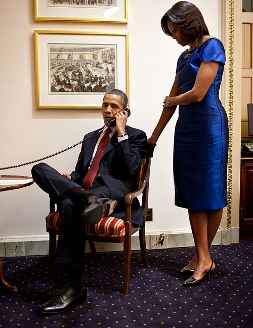 First Lady Michelle Obama stands behind the President as he makes a phone call