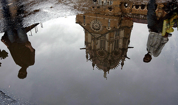 Reflections of pedestrians are seen in a puddle of rainwater outside the Chhatrapati Shivaji Terminus