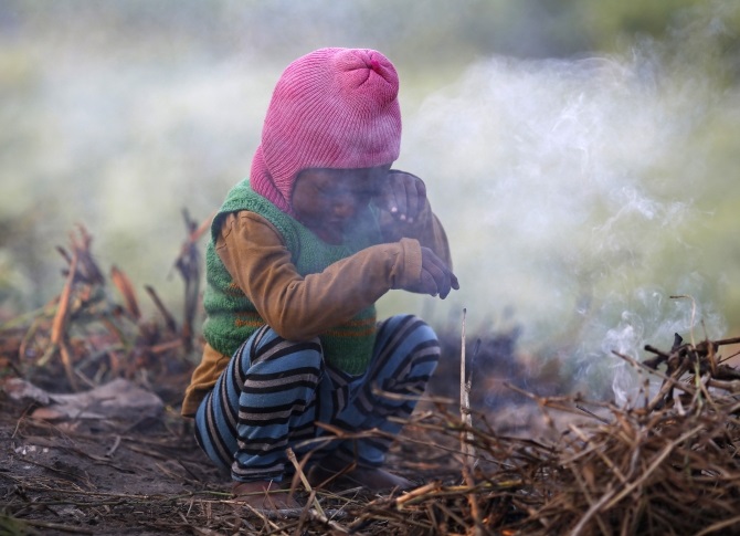 A boy wipes his eye as he sits near twigs which were set on fire by his parents to warm themselves at a vegetable field