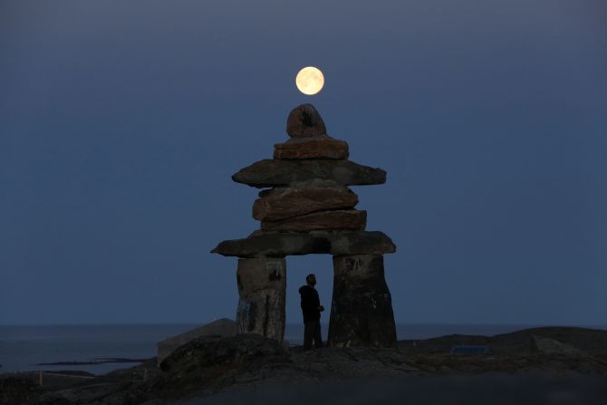 A man looks at a giant inukshuk as the moon rises above it in Rankin Inlet, Nunavut