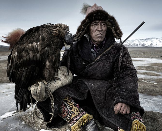 A hunter rests in his little house on the plains in western mongolia with his Eagle which is hooded and cat under the table!