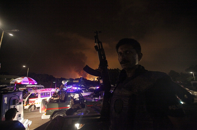 A policeman with a gun stands guard on a vehicle as smoke billows from Jinnah International Airport in Karachi