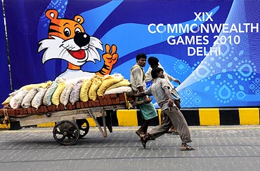 Labourers pull a handcart loaded with bricks and sand in front of boards advertising the Commonwealth Games.