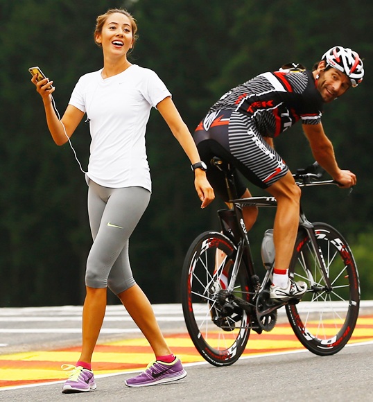 Mark Webber of Red Bull Racing rides his bike past Jenson Buttons girlfriend Jessica Michibata