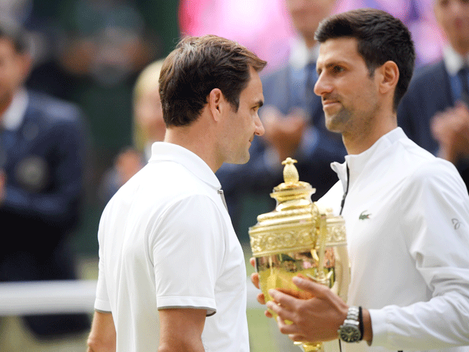 Roger Federer walks past Novak Djokovic who poses with the trophy after winning the 2019 Wimbledon Championships
